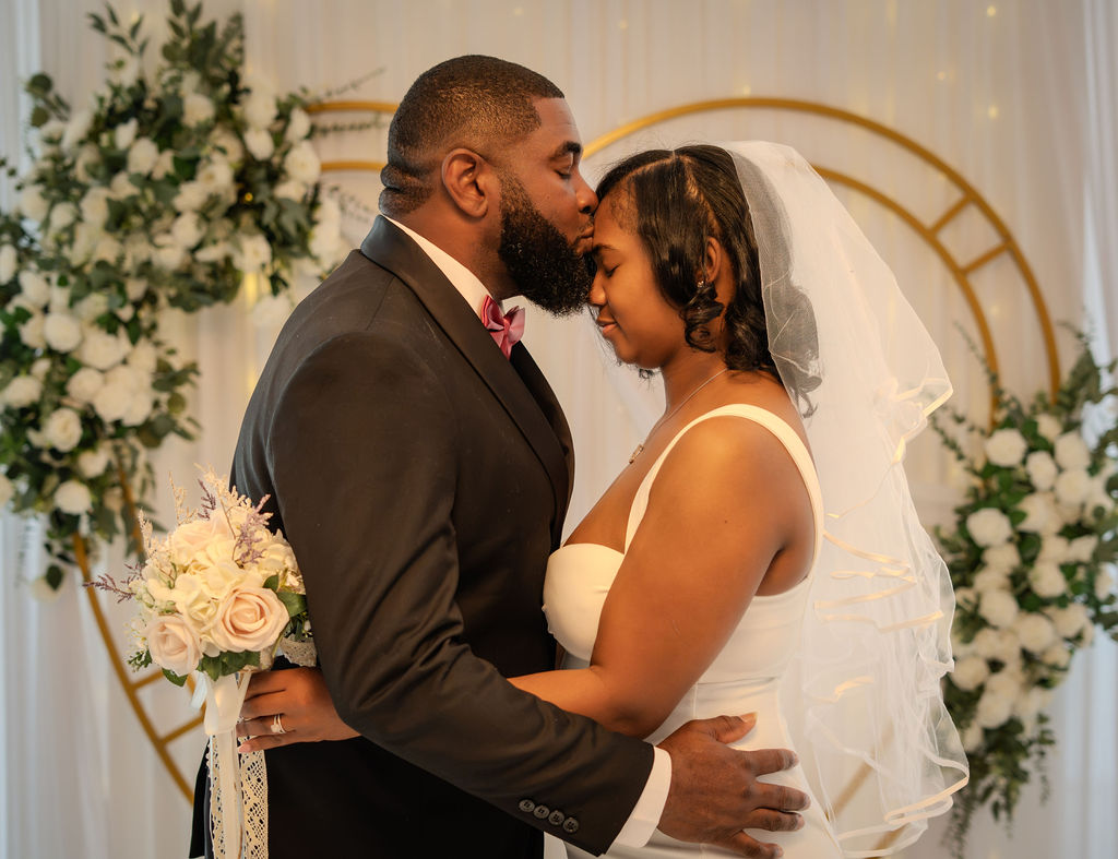 Groom kissing his bride's forehead in front of a gold arch with white roses